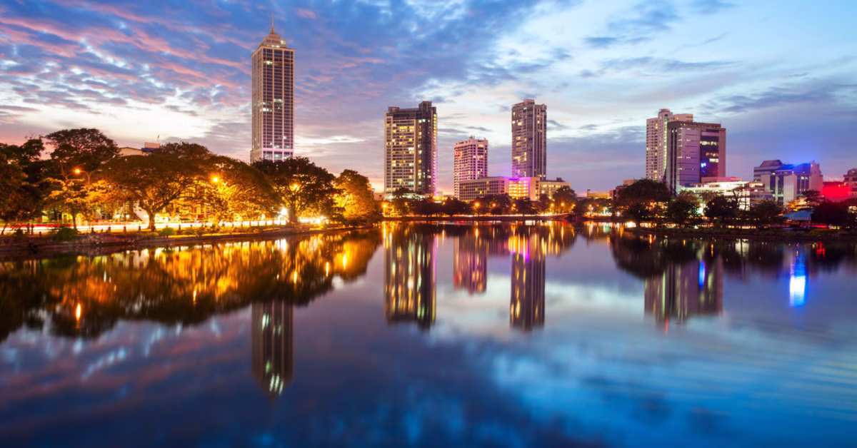 A city skyline with tall buildings reflecting in a lake at dusk by trees and colorful lights